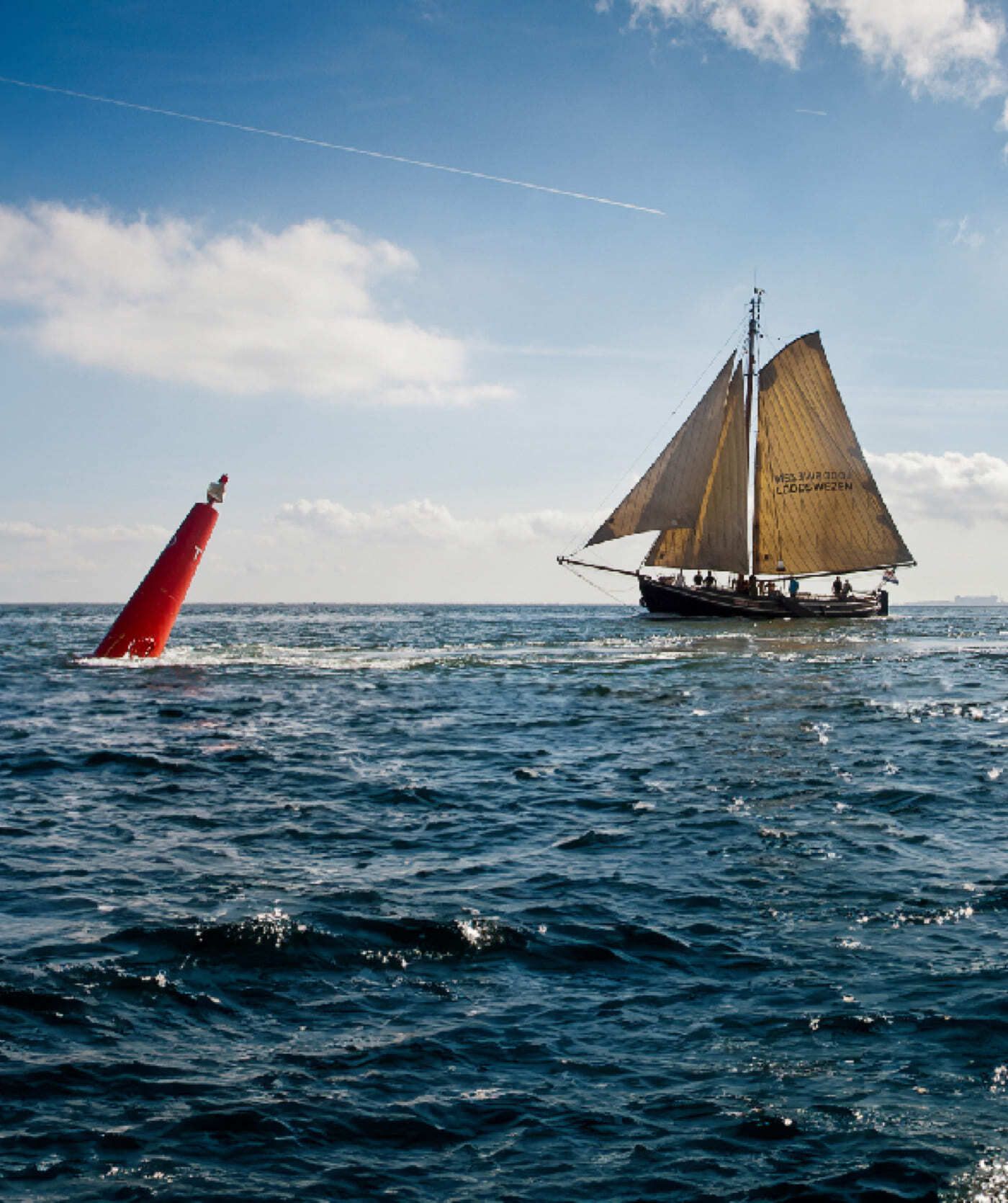 Een zeilboot met bruine zeilen vaart langs een gekantelde rode boei op open zee, onder een blauwe lucht met hier en daar een wolkje. De perfecte inspiratie voor creatieve websites of webshops.