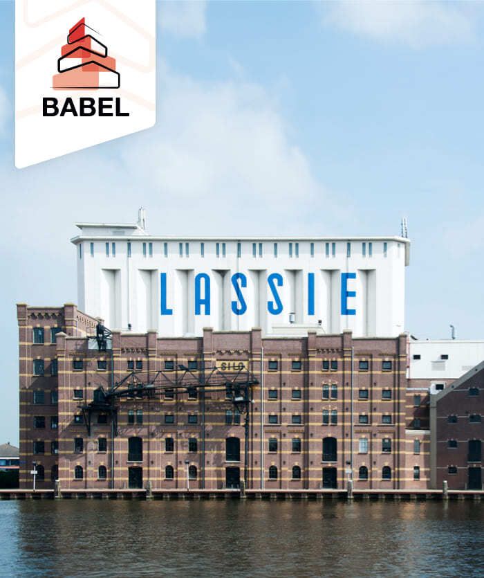 Een groot bakstenen gebouw met "LASSIE" in blauwe letters op het bovenste witte gedeelte, aan het water onder een gedeeltelijk bewolkte hemel. Linksboven staat het logo van Babel, dat hun expertise op het gebied van websites en online diensten benadrukt.