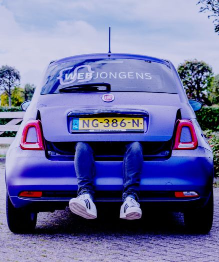 Een persoon zit in de open kofferbak van een paarse Fiat, met zijn benen naar buiten. De auto heeft een Nederlands kenteken en WEB JONGENS op de achterruit, wat wijst op hun expertise op het gebied van websites en online diensten.