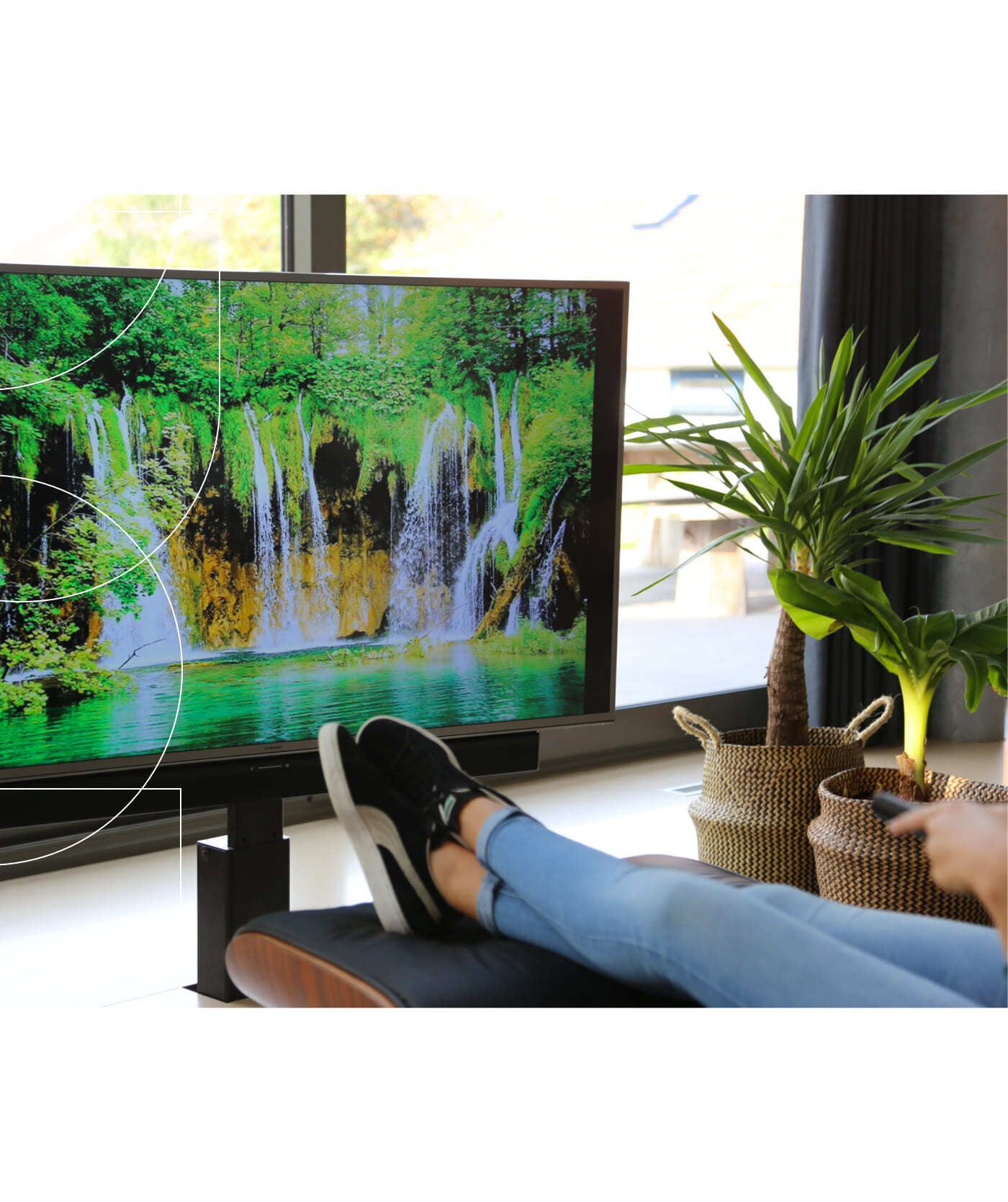 Een persoon met gekruiste benen zit achteroverleunend voor een tv waarop een waterval te zien is, en houdt een afstandsbediening vast. Naast de tv staan twee potplanten, die een rustig plekje creëren om op uw gemak websites of webshops te bekijken.