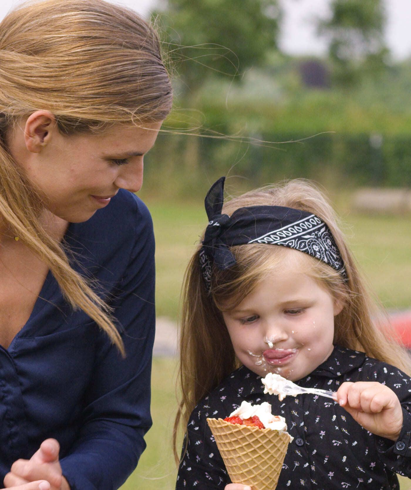 Een vrouw kijkt toe hoe een jong meisje met een bandana een ijsje uit een hoorntje eet met een lepel en er een beetje van op haar gezicht krijgt. Ze zijn buiten, met groen op de achtergrond, en genieten van een pauze na samen webshops te hebben bezocht.