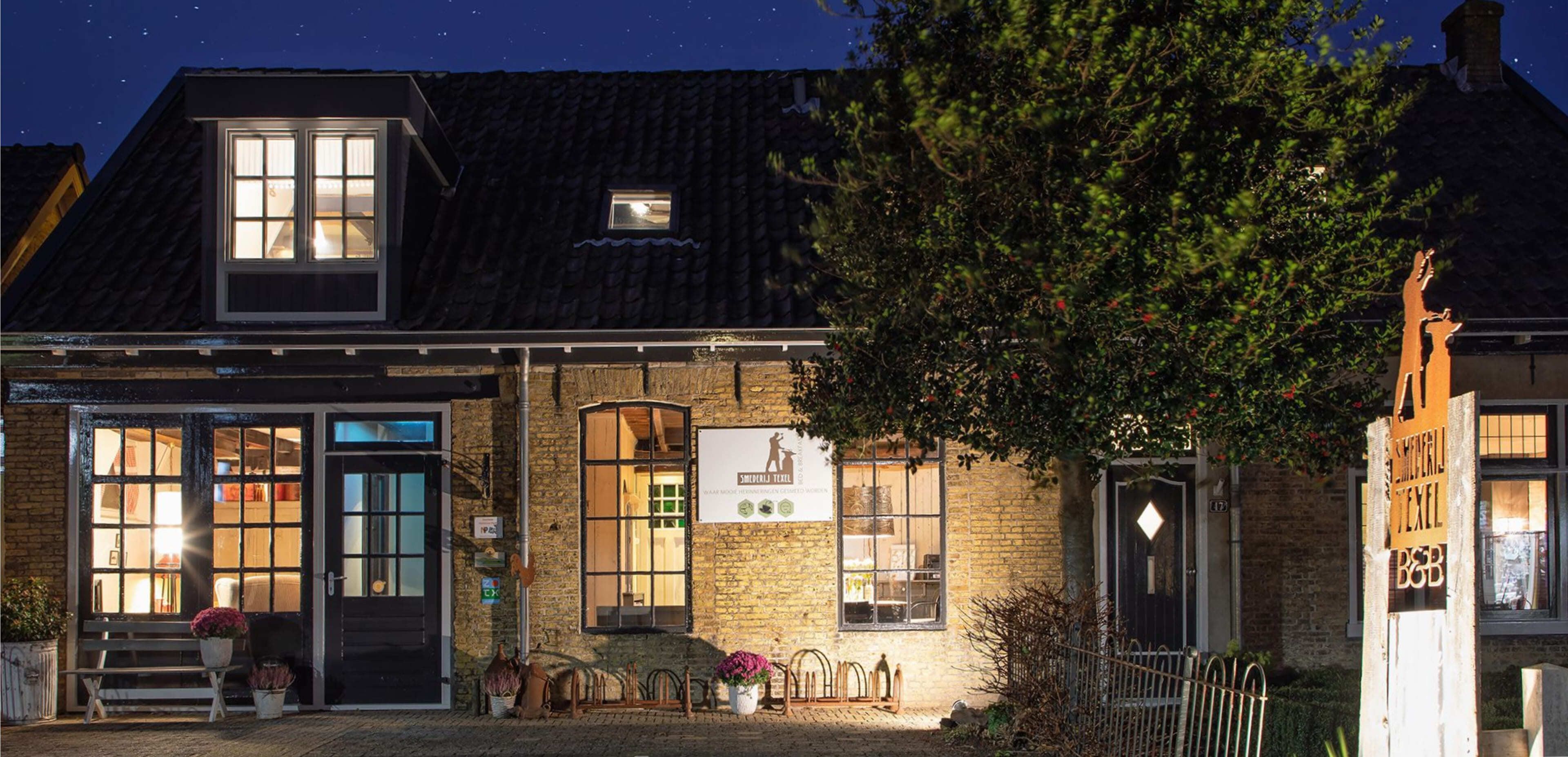 Een bakstenen gebouw met een zwart dak en 's avonds verlichte ramen, een grote boom aan de rechterkant en een houten bord met een paardenlogo op de voorkant: een gezellige plek die inspiratie kan bieden voor charmante webshops of creatieve websites.