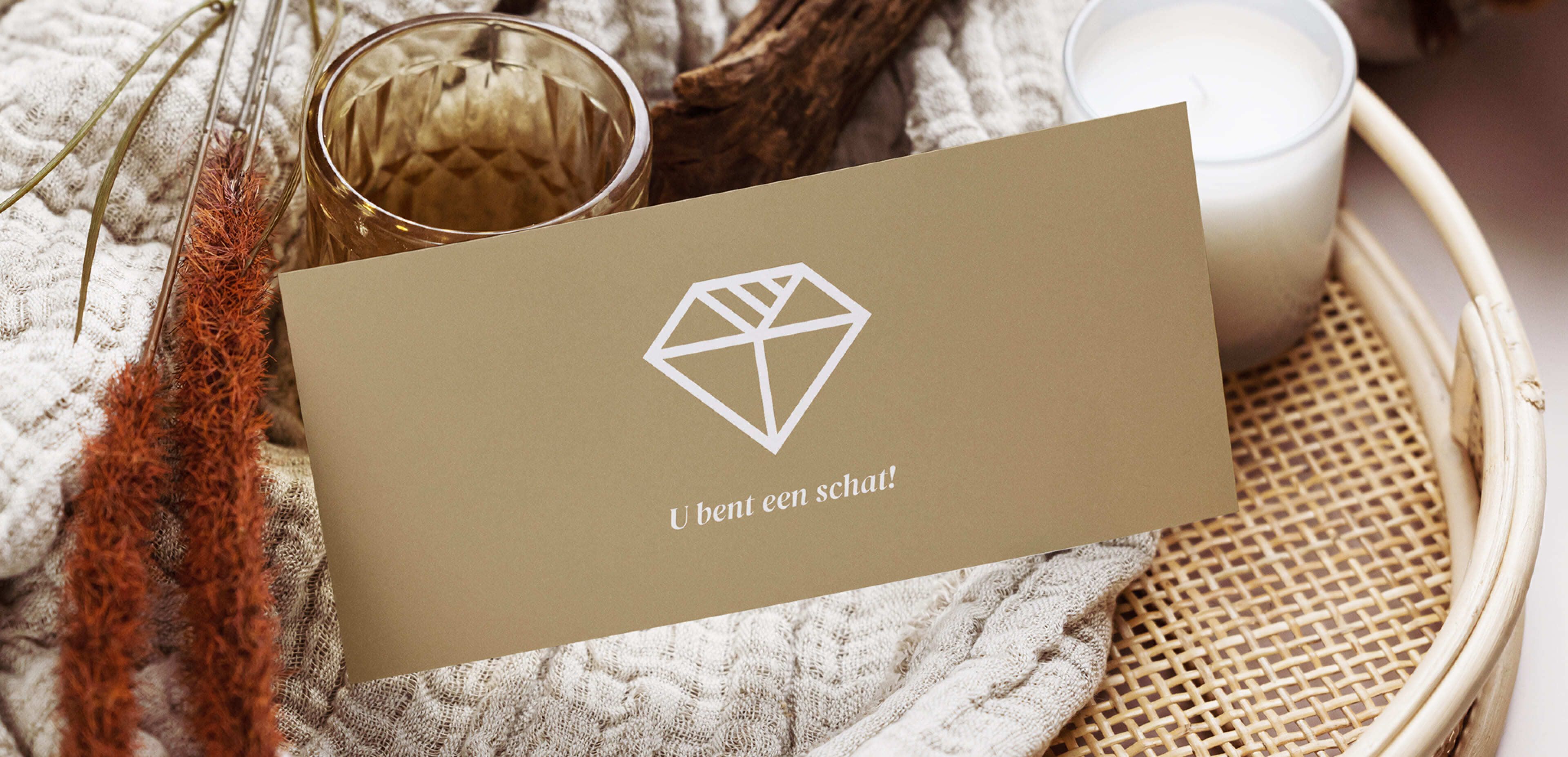 Een beige kaart met een witte ruit en de tekst "U bent een schat!", gelegd op een dienblad met een kaars, glas en geweven textiel. Ideaal voor webshops of online diensten die een persoonlijk tintje willen geven aan hun producten.