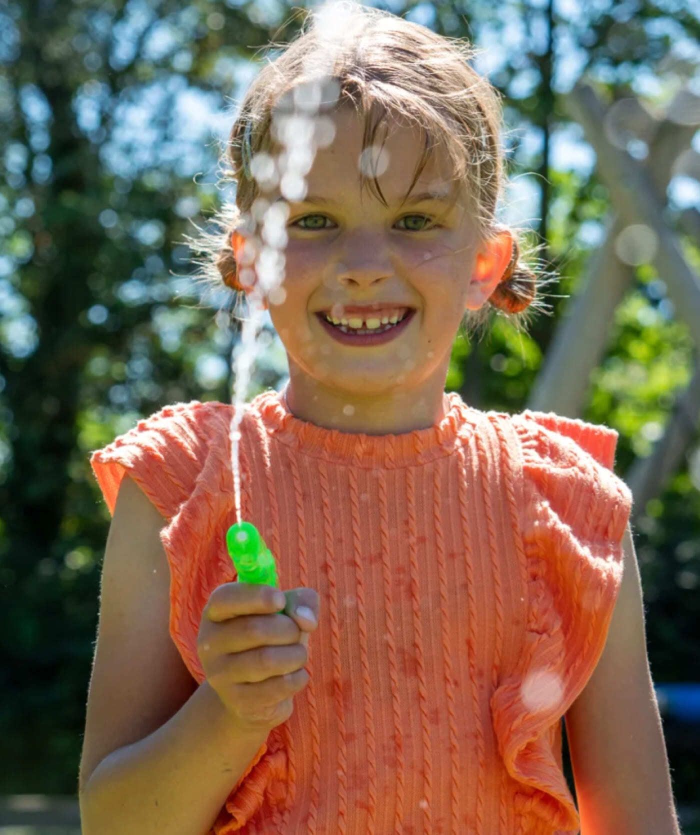 Een kind in een oranje shirt lacht terwijl hij op een zonnige dag buiten met een groen waterpistool water spuit. Het kind geniet van een pauze tijdens het snuffelen op leuke webshops en websites.
