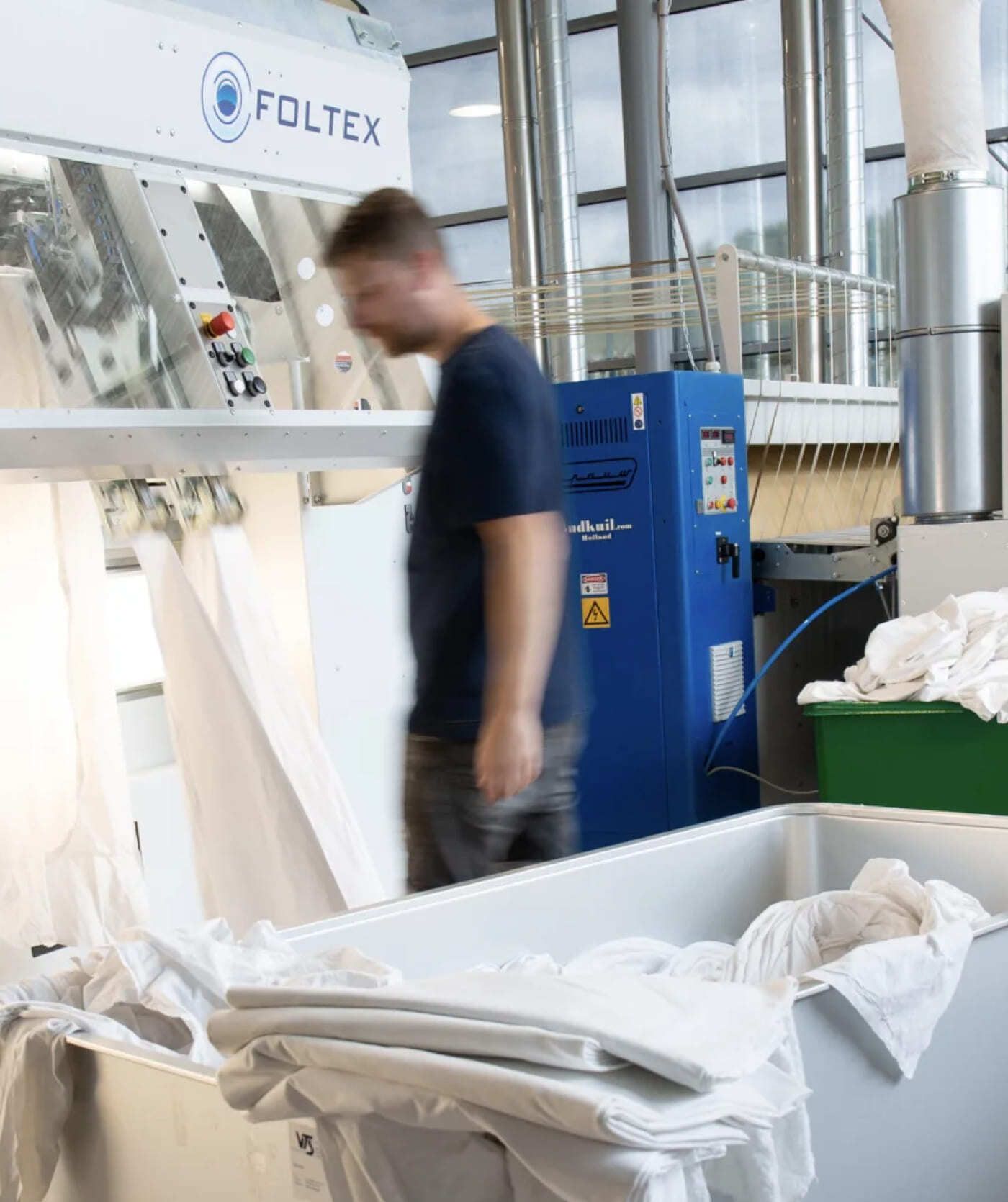 Een persoon staat bij een industriële wasmachine met wit linnengoed en bakken in een fabrieksomgeving en verwerkt benodigdheden voor webwinkels en onlinediensten.
